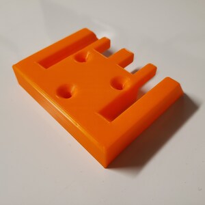 May include: Orange plastic 3D printed object with four slots and three holes.