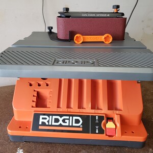 May include: A gray and orange belt sander with a stand. The stand has a storage compartment and a red switch. The sander is labeled "RIDGID".