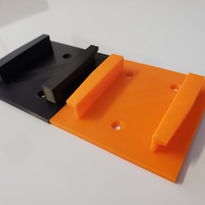 May include: Two 3D printed wall mounts, one black and one orange. The mounts have a rectangular shape with a raised lip on one side.