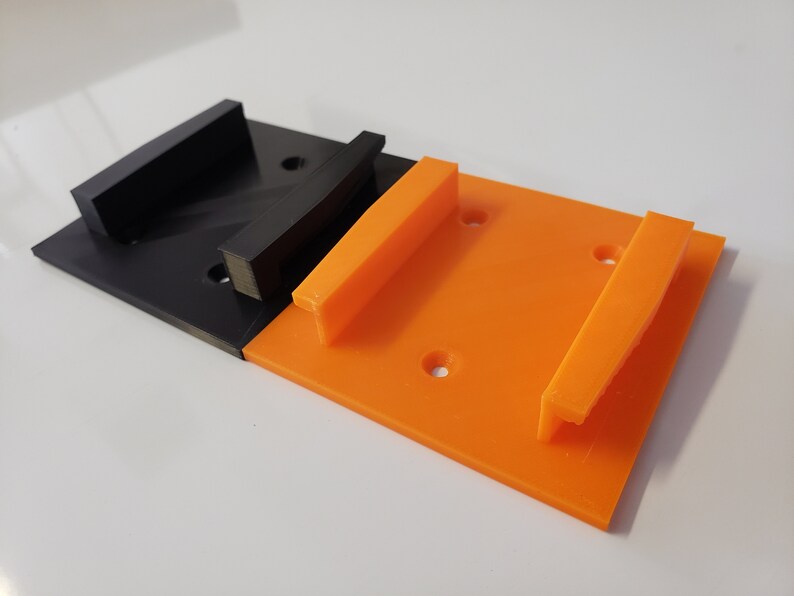 May include: Two 3D printed wall mounts, one black and one orange. The mounts have a rectangular shape with a raised lip on one side.