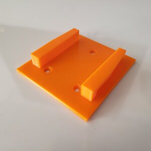 May include: Orange plastic wall mount with two slots for holding objects. The mount has four holes for attaching to a wall.