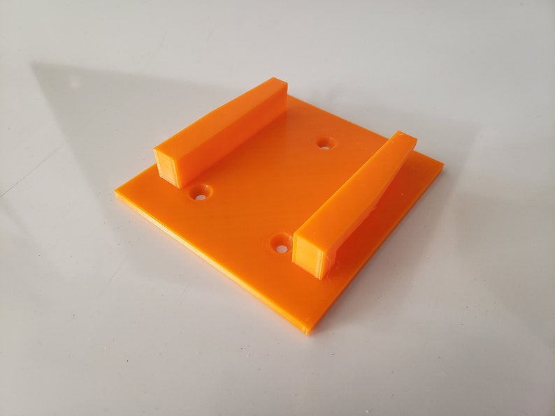 May include: Orange plastic wall mount with two slots for holding objects. The mount has four holes for attaching to a wall.