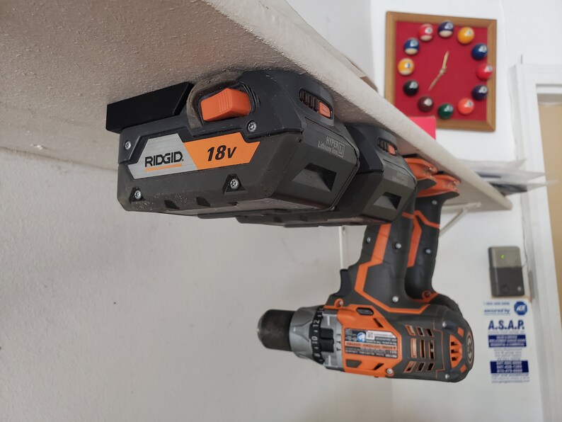 May include: Two black and orange 18V RIDGID batteries mounted on a shelf with a black and orange drill hanging below.
