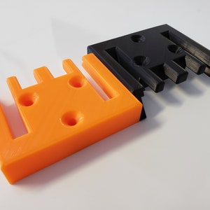 May include: Two 3D printed wall mounts, one orange and one black. Both mounts have a series of slots and holes for attaching items.