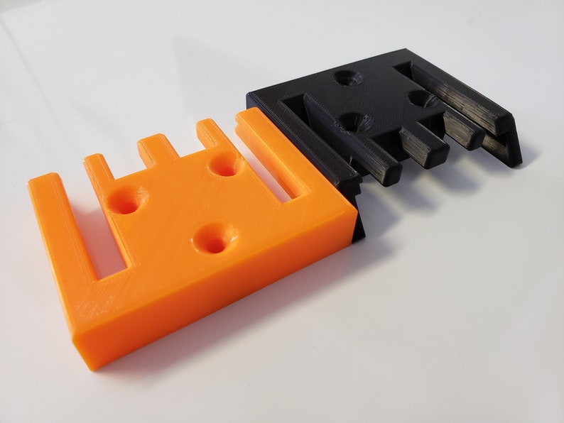 May include: Two 3D printed wall mounts, one orange and one black. Both mounts have a series of slots and holes for attaching items.