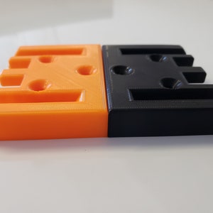 May include: Two 3D printed blocks, one orange and one black, with multiple holes and slots. The blocks are side by side.