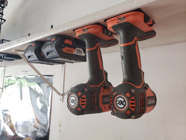 May include: Two black and orange cordless drills with a 4.0Ah battery hanging on a white shelf. The drills have a black and orange handle and a black and orange trigger. The drills are labeled "RIDGID" and "DXID".