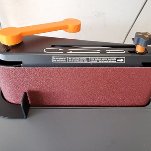 May include: A black belt sander with an orange handle and a black knob. The sander has a red sanding belt and a black base. The sander has text in English, Spanish, and French that says "Do not sand on this end of belt", "No use este extremo de la banda para lijar", and "Il ne faut pas poncer avec cette extremit de la bande abrasive".