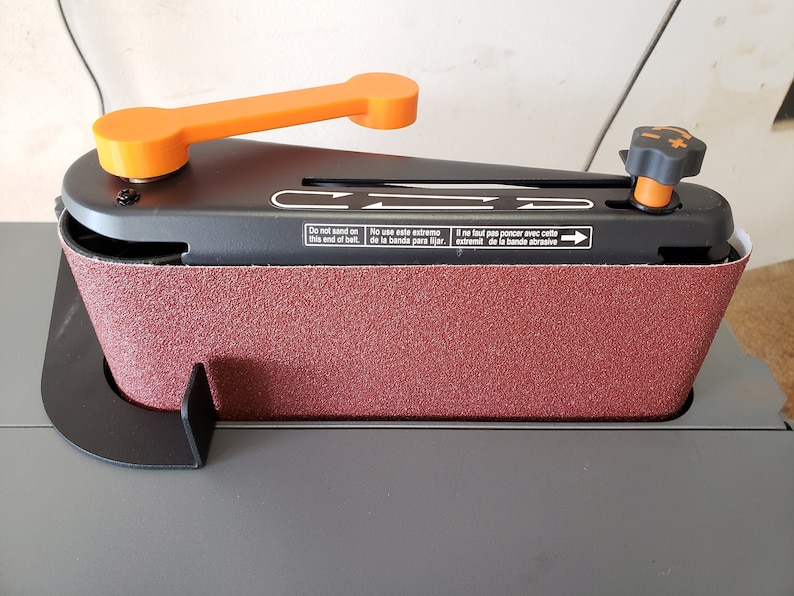 May include: A black belt sander with an orange handle and a black knob. The sander has a red sanding belt and a black base. The sander has text in English, Spanish, and French that says "Do not sand on this end of belt", "No use este extremo de la banda para lijar", and "Il ne faut pas poncer avec cette extremit de la bande abrasive".