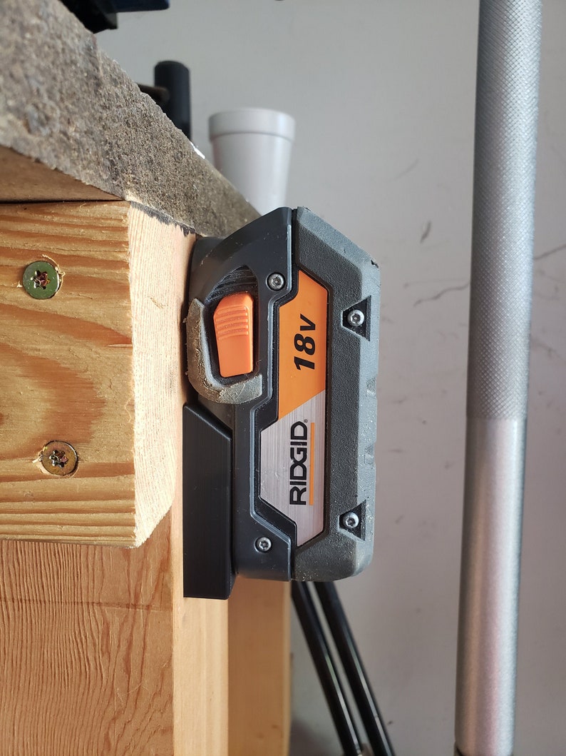 May include: A black and orange 18V RIDGID battery is mounted on a wooden surface.