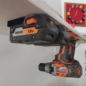 May include: Two black and orange 18V RIDGID batteries mounted on a shelf with a black and orange drill hanging below.