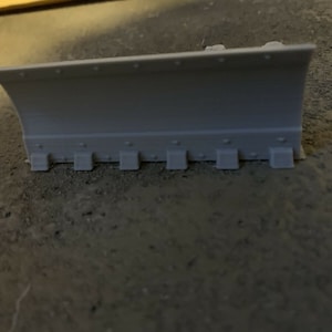 May include: A grey plastic snow plough blade with a curved front edge and a series of small rectangular attachments along the bottom.