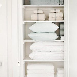May include: A white closet with multiple shelves, neatly organized with folded towels, pillows, and bedding. The items are in shades of white, cream, and light blue, creating a clean and minimalist aesthetic. The closet is well-lit, showcasing the organized storage.