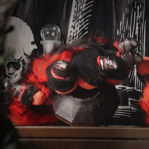 May include: A pair of furry paw gloves with red and black fur. The gloves have large, rounded red pads and black claws. The background includes a black and white patterned fabric and a skull.