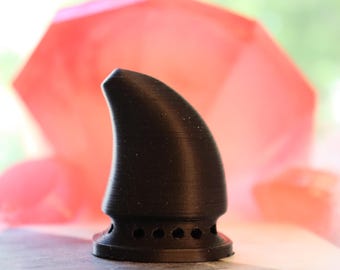 May include: A black, horn-shaped object with a rounded tip and a circular base featuring several small holes. The object is set against a blurred, pinkish-red background, suggesting a decorative or artistic piece.