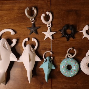 Peut inclure: Une collection de boucles d'oreilles faites à la main de différentes formes, dont des requins, des étoiles, des beignets et des anneaux à pointes. Les boucles d'oreilles sont blanches, noires et bleues, avec un crochet argenté. Les articles sont exposés sur une surface en bois.