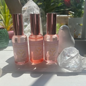 May include: Three pink glass spray bottles with rose gold lids are displayed. Each bottle has a label with text and a logo. A clear crystal and other decorative items are in the background, creating a natural aesthetic.