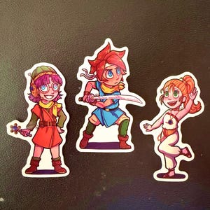 May include: Three cartoon stickers featuring characters with vibrant colors. One character wears a red outfit and glasses, another holds a sword and wears blue, and the third is in white and orange. The stickers have a fantasy theme.