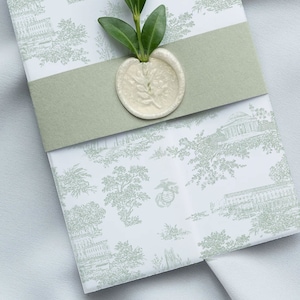 May include: A white envelope with a green ribbon and a wax seal. The envelope has a delicate pattern of green trees and buildings. A sprig of green leaves is placed on top of the ribbon.