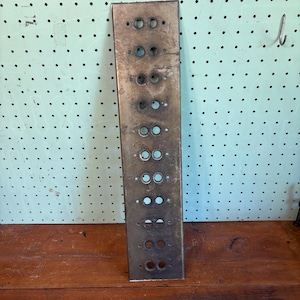May include: A rectangular, aged metal panel with multiple pairs of circular holes. The panel has a weathered appearance, with a slightly tarnished, bronze-like color. The holes are evenly spaced, suggesting a functional, possibly industrial, design.