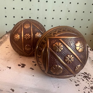May include: Two antique bronze door knobs with intricate floral and geometric patterns. The knobs have a round shape and a warm, metallic finish. They are displayed on a distressed white surface.
