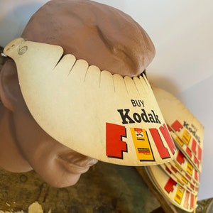 May include: A white cardboard sun visor with the text "BUY Kodak FILM" in red and black letters. The visor is on a mannequin head. There are several stacks of Kodak film boxes in the background.
