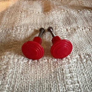 May include: Two bright red cabinet knobs with a ridged design. The knobs have a rounded shape and are attached to metal screws. The knobs are set against a textured, off-white fabric background.