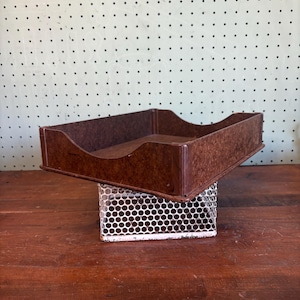 May include: A vintage brown metal desk tray with a textured finish, resting on a white wire mesh stand. The tray has a rectangular shape with curved sides. The background features a pegboard wall.