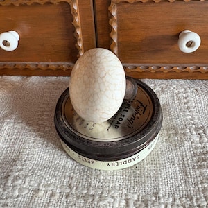 May include: A cream-colored, egg-shaped object with a cracked pattern sits atop a round, dark metal tin. The tin has a white label with black text, including the words "SADDLERY" and "APPLE SOAP". The background includes a wooden drawer and a textured, off-white surface.
