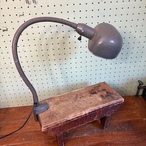 May include: A gray metal desk lamp with a flexible gooseneck arm and a dome-shaped shade, featuring a clamp base attached to a small, rectangular, weathered wooden bench with four short legs and a dark, worn finish.
