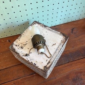 May include: An antique oil lamp with a white glass bulb and a brass and metal base. The lamp sits on a square, distressed white box with a woven edge. The background is a wooden surface and a mint green pegboard.