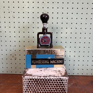 May include: An antique Bates Numbering Machine with a black handle and a red and black circular label. The machine sits atop a box with the word "Bates" and "Numbering Machine" printed on it. The box is on a white metal perforated container.