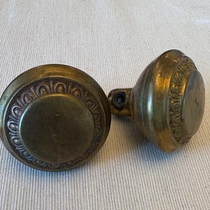 May include: Two antique brass door knobs. One knob is round with a decorative circular pattern around the edge. The other knob is oval-shaped with a similar decorative pattern. Both knobs have a tarnished, aged appearance.
