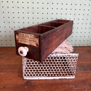 May include: A vintage wooden drawer with three compartments, a white ceramic knob, and a metal label that reads "WB-166 T/II CARBOLOX TIPPED WAUKESHA REAPER BLADES." The drawer sits atop a white metal perforated box.
