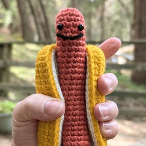 May include: A handmade, crocheted hot dog toy with a smiling face. The hot dog is a reddish-orange color, nestled in a yellow bun with white and black accents. Black feet complete the whimsical design.