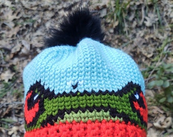 May include: A blue and red crocheted beanie with a black pom pom on top. The beanie has a green, black, and red design that resembles a frog.