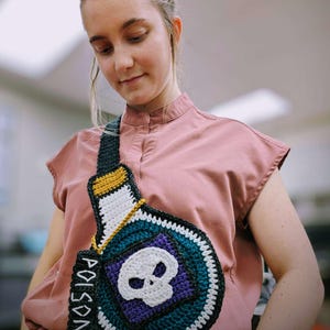 Puede incluir: Bolso bandolera de ganchillo hecho a mano con forma de botella de veneno. El bolso presenta una calavera blanca sobre un cuadrado morado, con la palabra "POISON" en una etiqueta. El bolso tiene una combinación de colores negro, verde azulado, blanco, amarillo y dorado.
