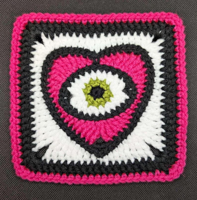 May include: A crocheted square with a pink heart design. The heart has a white and black eye with a green center.