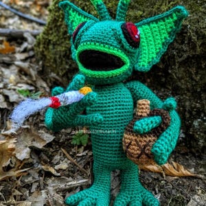 Può includere: Una creatura verde all'uncinetto fatta a mano con grandi orecchie, occhi rossi e una bocca spalancata. Tiene in mano una sigaretta accesa e una borsa a forma di ghianda marrone e beige. La creatura si trova in un ambiente naturale all'aperto.