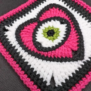May include: A crocheted square with a pink heart design in the center. The heart is surrounded by white and black crocheted circles. The outer border of the square is pink.