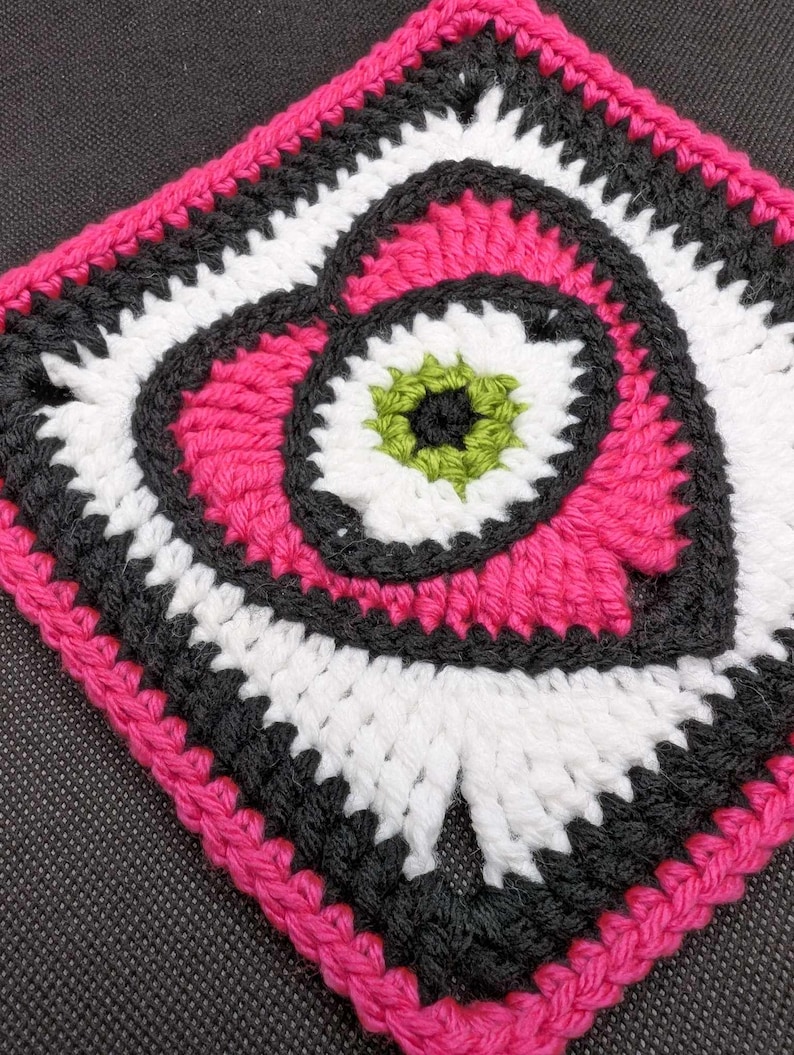May include: A crocheted square with a pink heart design in the center. The heart is surrounded by white and black crocheted circles. The outer border of the square is pink.