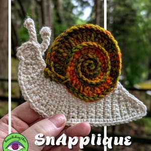 May include: A crochet snail applique featuring a cream-colored body with two antennae and a spiraled shell in variegated shades of orange, green, and brown. The text SnApplique is printed in white at the bottom of the image.