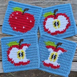 Op de afbeelding: Vier gehaakte vierkanten met verschillende appelontwerpen. Elk vierkant is blauw met een rood, wit en groen appelontwerp.