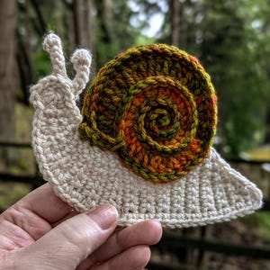 Könnte beinhalten: Gehäkelte Schneckenapplikation mit cremefarbenem Körper und einer spiralförmigen Schale in Orange-, Grün- und Brauntönen. Die Schnecke hat zwei cremefarbene Fühler. Das handgefertigte Kunsthandwerk wird vor einem verschwommenen natürlichen Hintergrund gehalten.