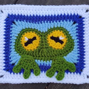 May include: A crocheted square featuring a green owl with yellow eyes and a blue and white border.