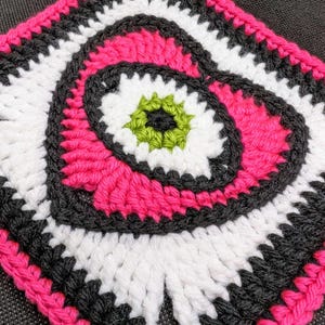 May include: A crocheted square with a pink, white, black, and green design. The center of the square features a spiral pattern.