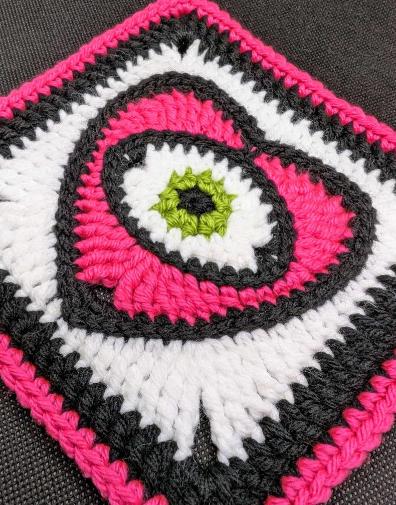 May include: A crocheted square with a pink, white, black, and green design. The center of the square features a spiral pattern.