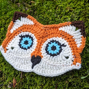 Puede incluir: Una máscara de zorro de crochet hecha con hilo blanco, naranja y marrón. La máscara tiene ojos azules y una nariz negra.