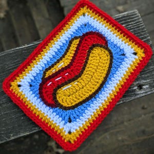 May include: A handmade crochet hot dog coaster. The coaster features a red, yellow, blue, and white striped border, with a red hot dog and yellow bun in the center. The coaster is square and sits on a wooden surface.