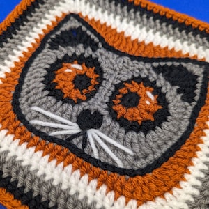 May include: Crocheted square with a grey cat face in the centre. The cat has black eyes, a black nose, and white whiskers. The square is made with orange, white, grey, and black yarn.